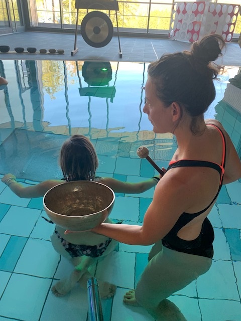 Une femme guide une enfant dans une séance de bols chantants dans une piscine.